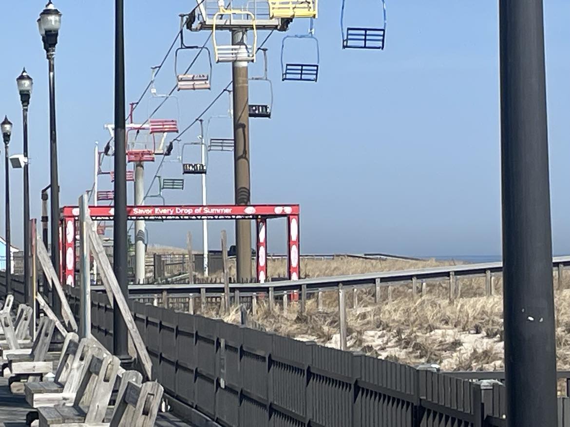"Savor Every Drop of Summer" written on an amusement park ride. There's a tram line running above it, the scene is at the beach, water is distant in the background